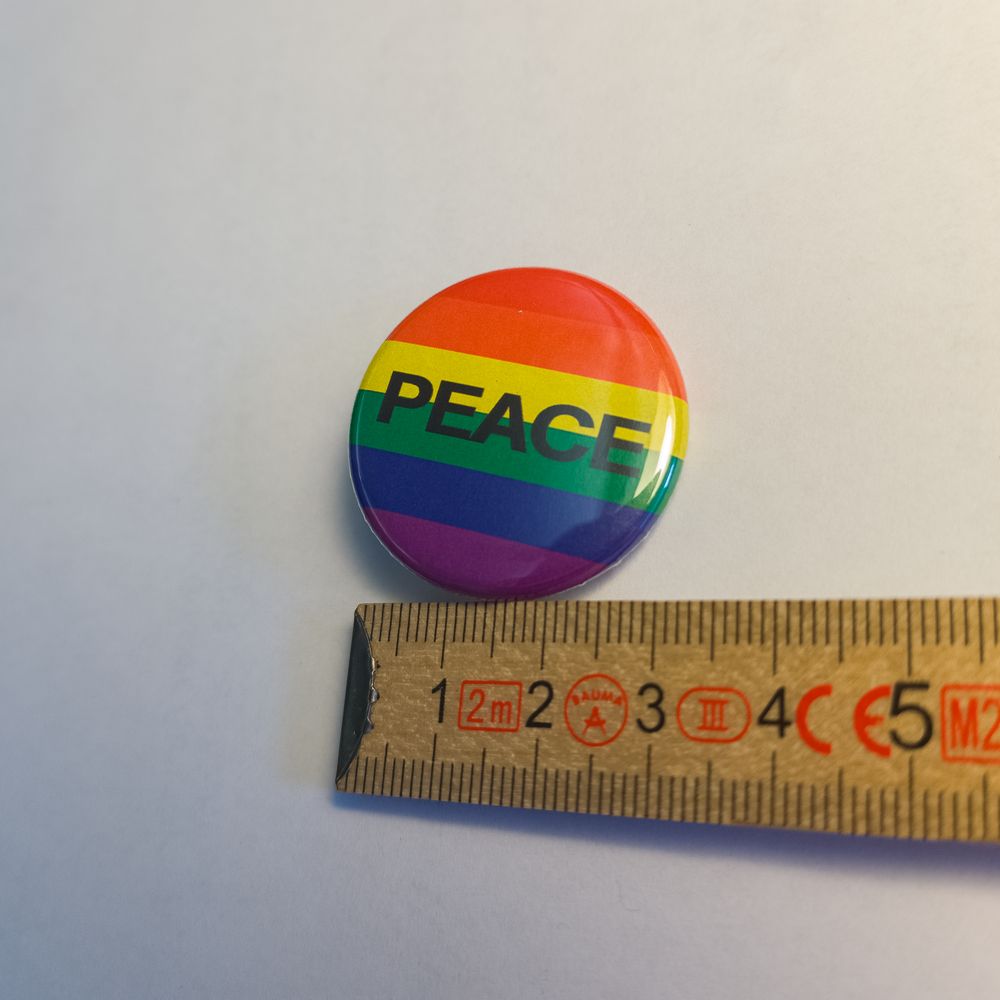 Runder Metall-Button mit klassischem Regenbogen-Hintergrund. Quer über die Mitte ist in klaren, schwarzen Großbuchstaben das Wort "PEACE" gedruckt. Der Anstecker liegt an einem Zollstock und misst 31 Millimeter.