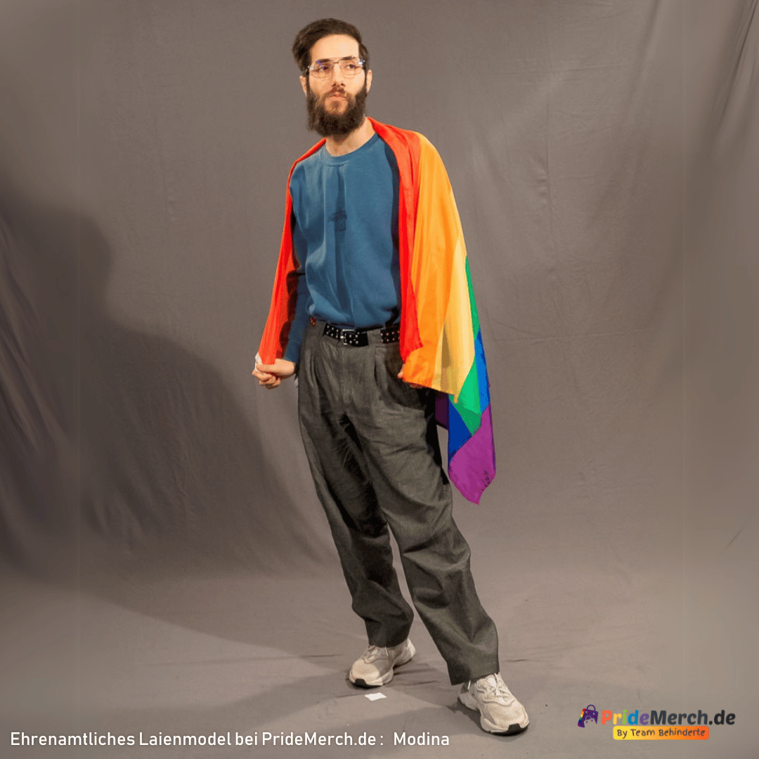 Unser ehrenamtliches Laienmodell Modina steht vor einem grauen Fotohintergrund. Er hat dunkle Haare, einen dichten Bart und trägt eine feine Brille. Bekleidet ist er mit einem blauen Pullover, einer grauen Anzughose und weißen Sneakern. Er hat sich die sechsstreifige Regenbogenflagge wie einen bunten Umhang über seine linke Schulter gelegt und blickt entspannt zur Seite.