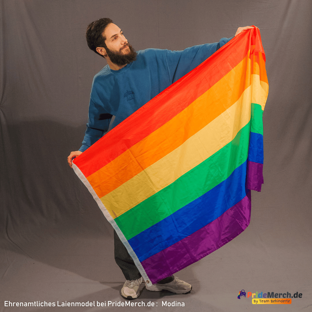 Laienmodell Modina steht frontal vor dem grauen Fotohintergrund und breitet die Regenbogenflagge mit beiden Händen seitlich neben seinem Körper aus. Er schaut nach oben rechts in Richtung der Flagge. Am unteren linken Rand der Fahne ist deutlich die weiße Stoffkante mit einer kreisrunden, silbernen Metallöse zur Befestigung zu erkennen.