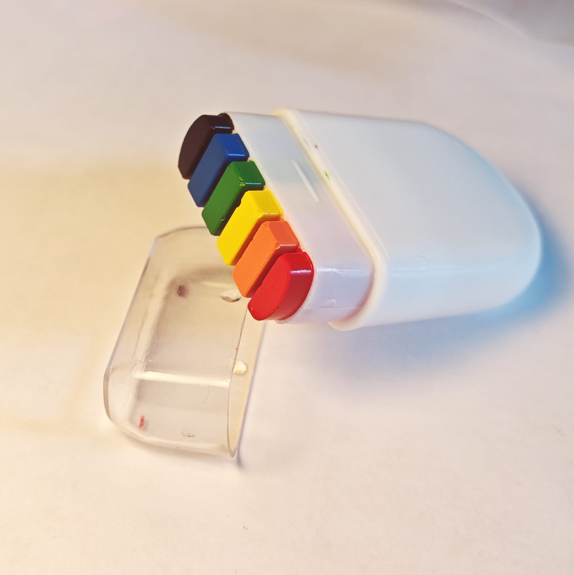 Ein weißer, abgerundeter Schminkstift liegt geöffnet auf einer hellen Unterlage. Aus der oberen Öffnung ragen sechs feste Farbblöcke in Schwarz, Blau, Grün, Gelb, Orange und Rot heraus. Die transparente Kunststoffkappe wurde abgenommen und liegt schräg unter dem Stift, bereit, wieder aufgesteckt zu werden.
