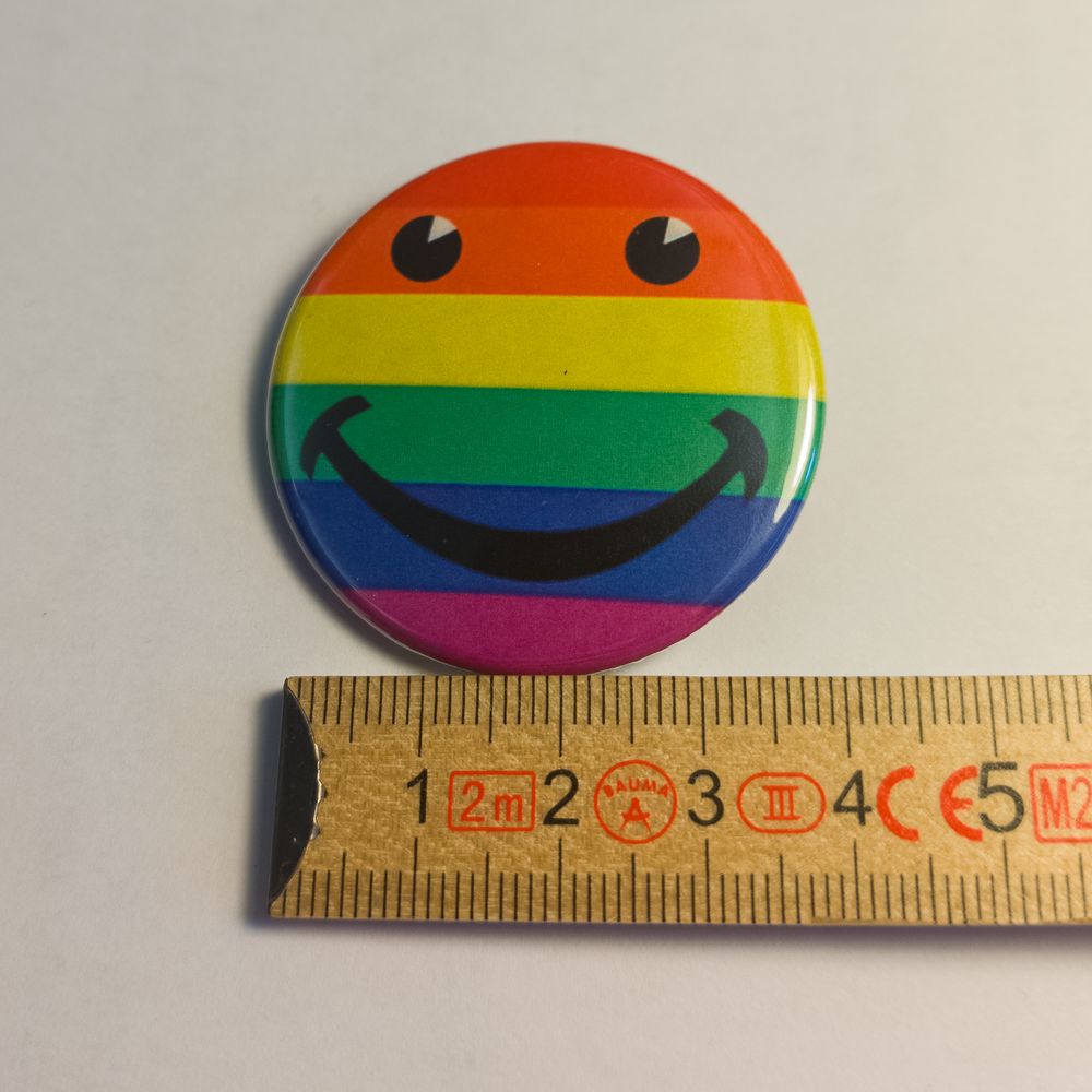 Ein runder Metall-Button. Auf dem bunten Hintergrund der sechsstreifigen Regenbogenflagge ist ein fröhliches Smiley-Gesicht mit schwarzen Augen und einem breiten Lächeln aufgedruckt. Neben dem Pin liegt ein Zollstock, der 37 Millimeter Durchmesser zeigt.