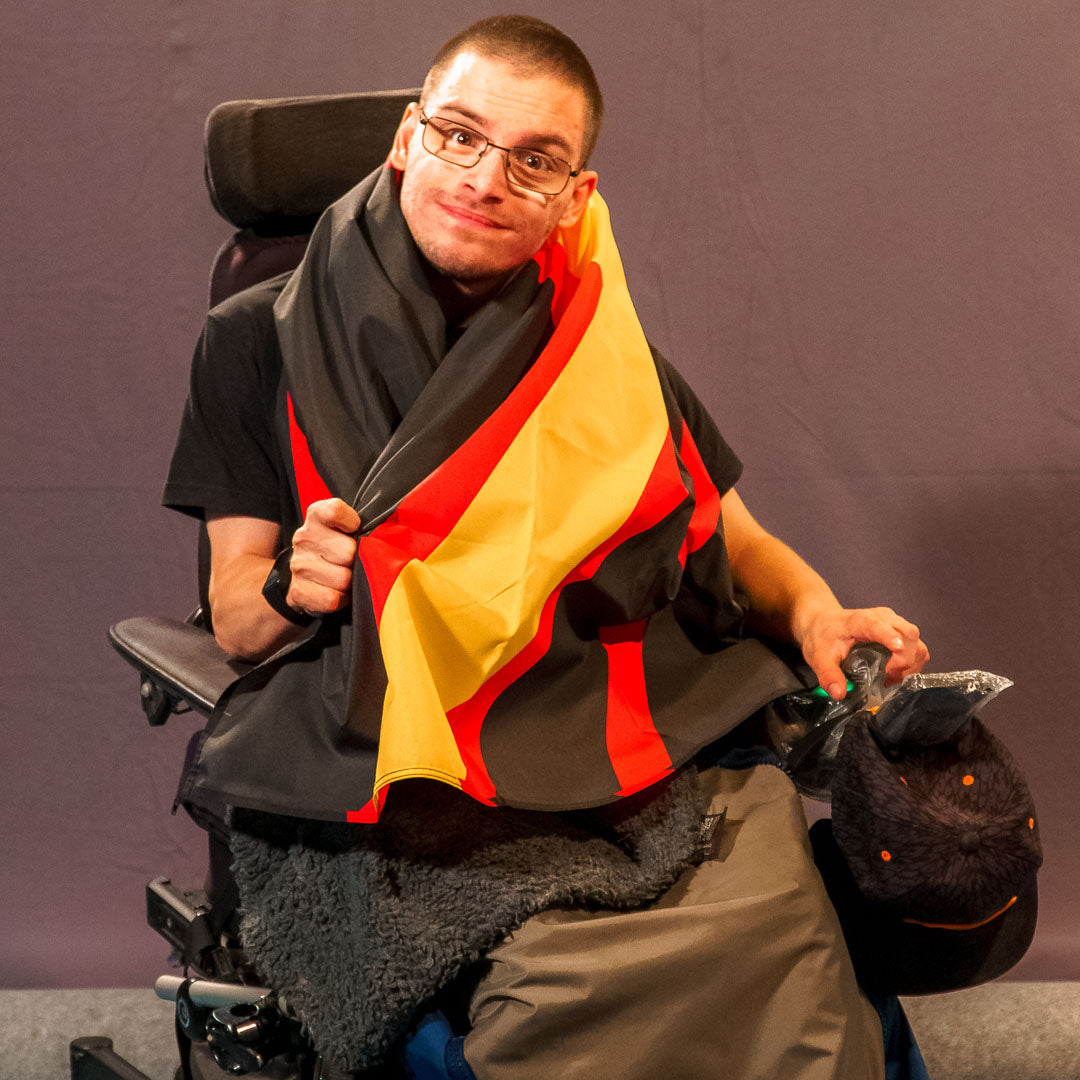 Sandro Weigang lächelt freundlich in die Kamera. Er sitzt im Rollstuhl und hat sich die Rubber Pride Flagge eng wie einen schützenden Umhang um die Schultern und den Oberkörper gelegt. Der tiefschwarze Stoff und die leuchtend gelben und roten Linien des markanten Winkels kommen durch den weichen Faltenwurf hervorragend zur Geltung.