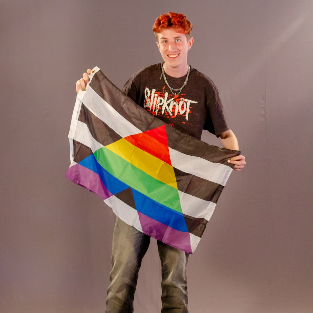 Das ehrenamtliche Laienmodell steht lachend vor einem grauen Fotohintergrund. Er hat kurze, rötlich gefärbte Haare, trägt ein schwarzes „Slipknot“-T-Shirt, mehrere silberne Halsketten und eine dunkelgraue Jeans. Mit beiden Händen hält er die schwarz-weiße Straight Ally Flagge mit dem bunten Regenbogen-A vor sich aufgefaltet.