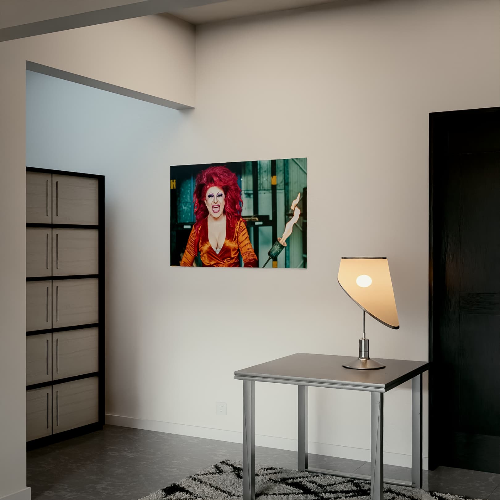 Ein Poster mit den Maßen 90x60 cm an einer hellen Wand in einem modern eingerichteten Raum. Das Poster zeigt Joyce DeLone mit leuchtend roten Haaren, dramatischem Make-up und einem orangefarbenen Outfit, während sie einen Flammenwerfer hält. Der Titel des Kunstwerks lautet „Flammenwerfer“. Im Vordergrund steht ein minimalistischer Tisch mit einer stilvollen Tischlampe, links ist ein schlichter Schrank zu sehen. Die Szene vermittelt eine moderne, künstlerische Atmosphäre