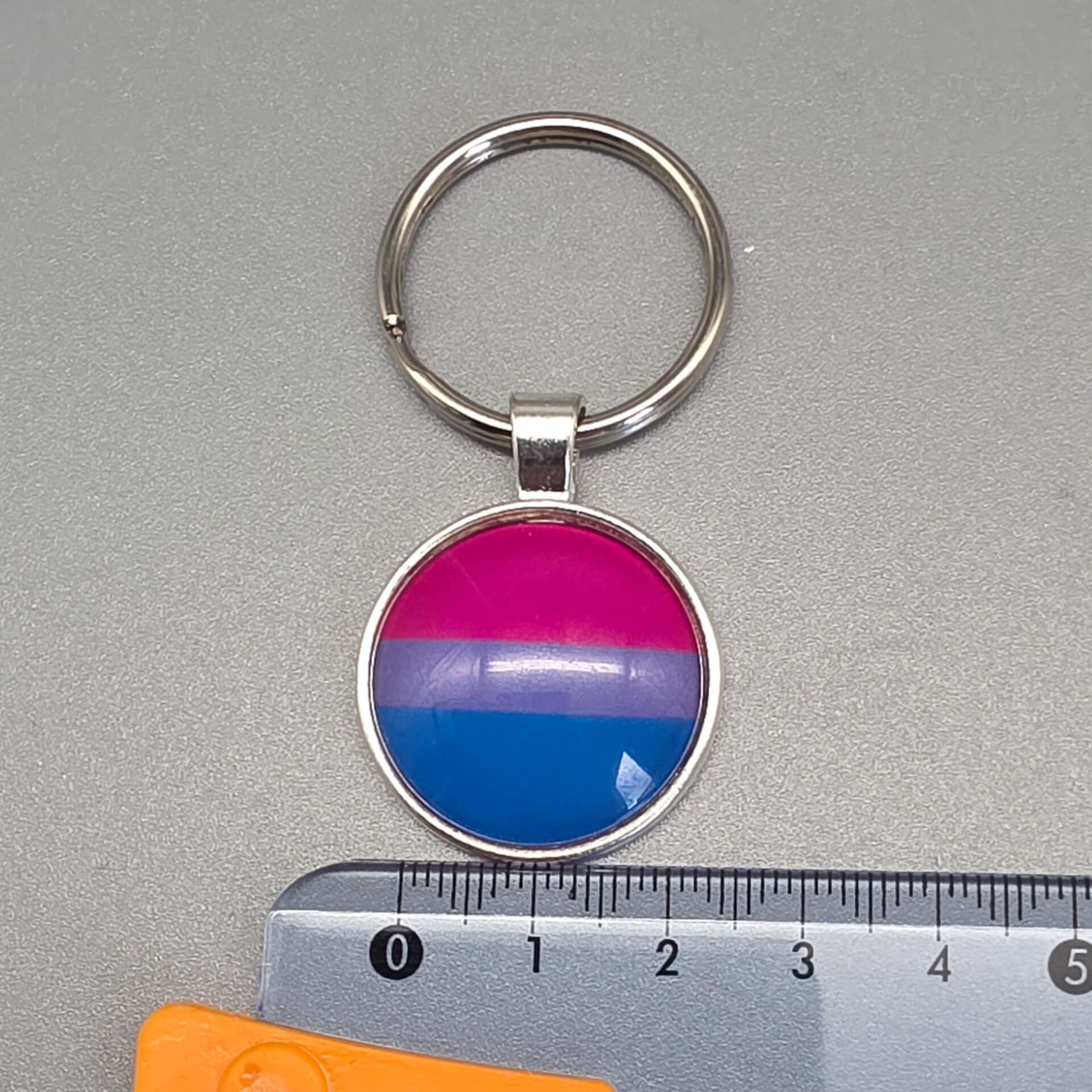 Bisexuell Pride Schlüsselanhänger in Silber mit Bi Flagge unter Glasdom, dargestellt neben einem Lineal zur Größenangabe von circa 25 mm Durchmesser.