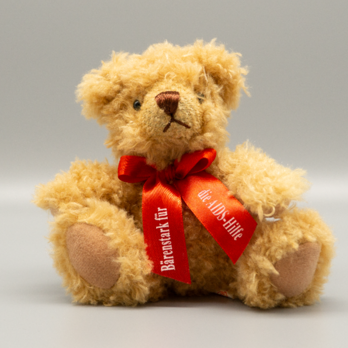 Ein goldbrauner Aids-Teddy aus dem Jahre 2011 mit besonders langem, wuscheligem Fell sitzt frontal vor der Kamera. Um den Hals ist eine rote Schleife mit dem deutlichen, weißen Aufdruck „Bärenstark für die AIDS-Hilfe“ fest gebunden. Die dunklen Knopfaugen heben sich stark von der helleren Schnauze ab.