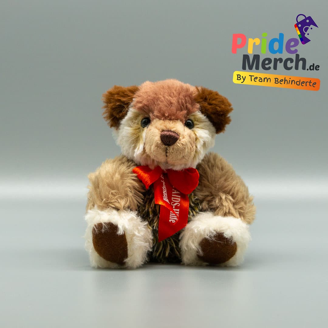 Ein sehr individueller Aids-Teddy mit mehrfarbigem, strubbeligem Fell: Der obere Teil des Kopfes ist dunkelbraun, die Schnauze weiß, und der Körper ist in verschiedenen Braun- und Weißtönen meliert. Die glänzende rote Schleife der AIDS-Hilfe sticht aus dem wuscheligen Plüschmuster leuchtend hervor.
