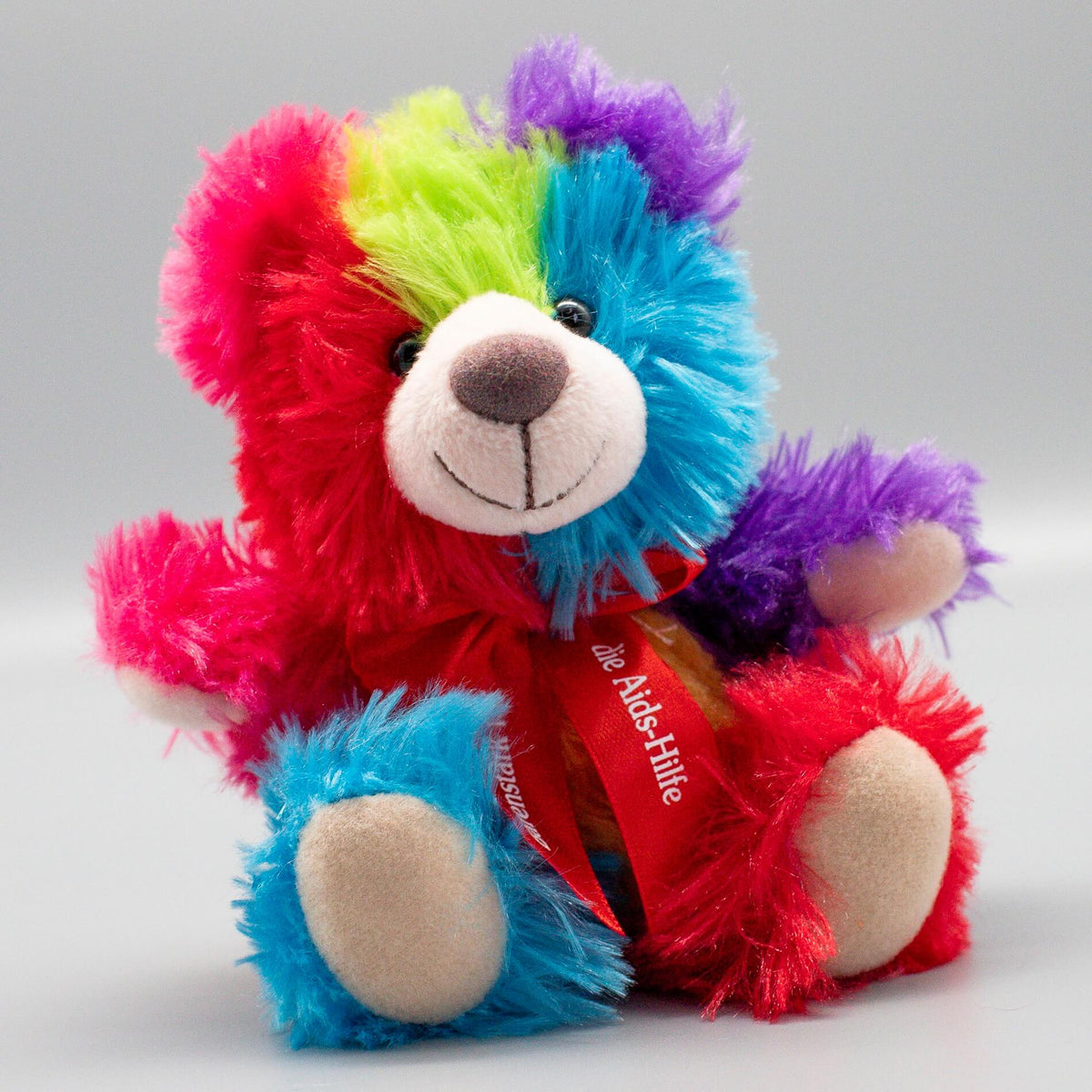 Farbenfroher Aids-Teddy aus dem Jahr 2025 mit leuchtendem Regenbogenfell. Die traditionelle rote Schleife um den Hals trägt den Schriftzug „die Aids-Hilfe“ und verbindet queere Sichtbarkeit direkt mit der Solidarität für HIV-positive Menschen.