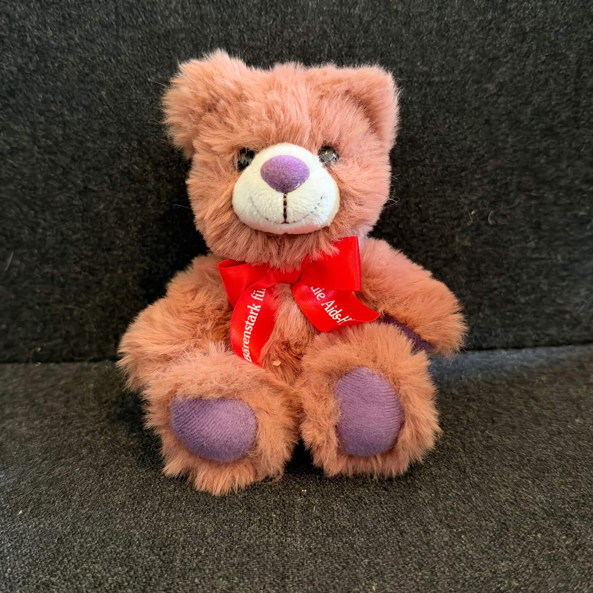 Flauschiger Plüsch-Teddy des aktuellen Jahrgangs 2026 in rotbrauner Farbe mit auffällig lila gefärbter Nase und lila Fußsohlen. Um den Hals ist eine leuchtend rote, glänzende Schleife mit dem weißen Schriftzug „Bärenstark für die Aids-Hilfe“ gebunden.