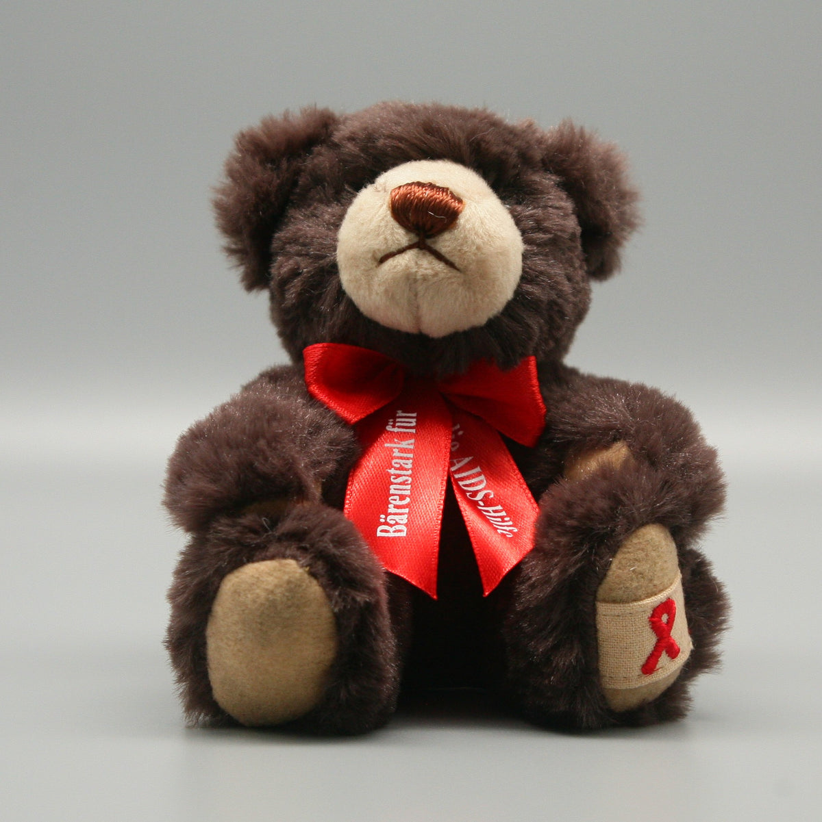 Klare Frontalansicht des dunkelbraunen Aids-Teddys. Das dunkle, dichte Fell bildet einen starken farblichen Kontrast zur leuchtend roten Schleife mit dem weißen Schriftzug „Bärenstark für die AIDS-Hilfe“. Auf dem linken, beigefarbenen Fußpad ist eine kleine rote Schleife eingestickt.