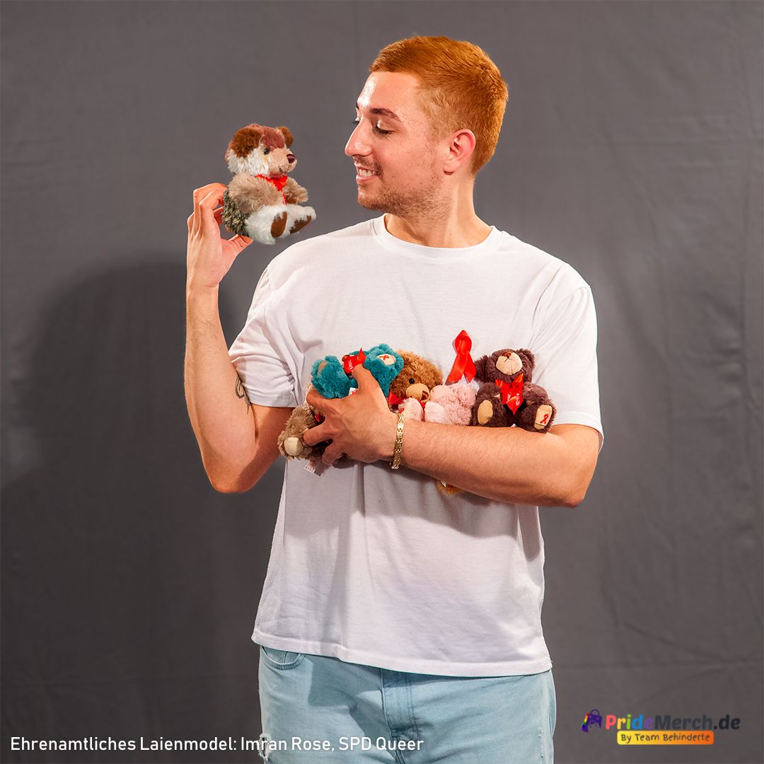 Das ehrenamtliche Laienmodel Imran Rose lächelt einen kleinen, braun-weißen Aids-Teddy an, den er in der rechten Hand auf Augenhöhe hält. Im linken Arm trägt er liebevoll und schützend eine ganze Sammlung bunter Teddys sowie eine große rote Schleife. Er trägt ein weißes T-Shirt vor grauem Hintergrund, was die bunten Farben der Bären stark hervorhebt.