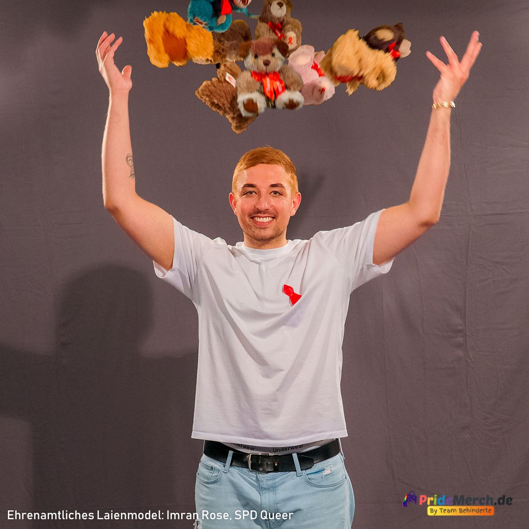 Mit einem strahlenden Lächeln und erhobenen Armen wirft das Model Imran Rose fröhlich eine Handvoll verschiedener, bunter Aids-Teddys in die Luft, die direkt über seinem Kopf schweben. An seinem weißen T-Shirt ist eine leuchtend rote Solidaritätsschleife angesteckt. Ein sehr dynamisches, lebensfrohes Bild vor grauem Hintergrund.