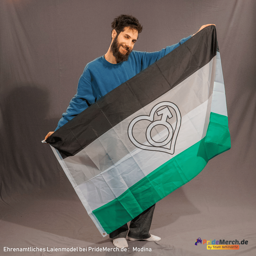In der Studioaufnahme präsentiert das ehrenamtliche Laienmodel Modina mit einem freundlichen Lächeln und vollem Bart die geöffnete Androphilie Pride Flagge vor grauem Studiohintergrund. Er hält die Pride Flagge im Format 150x90 cm schräg ausgebreitet, sodass die vier Querstreifen in Schwarz, Grau, Weiß und Grün sowie das markante weiße Herz mit dem Mars-Symbol im Zentrum perfekt zu sehen sind. Unten rechts befindet sich das Logo von PrideMerch.de.