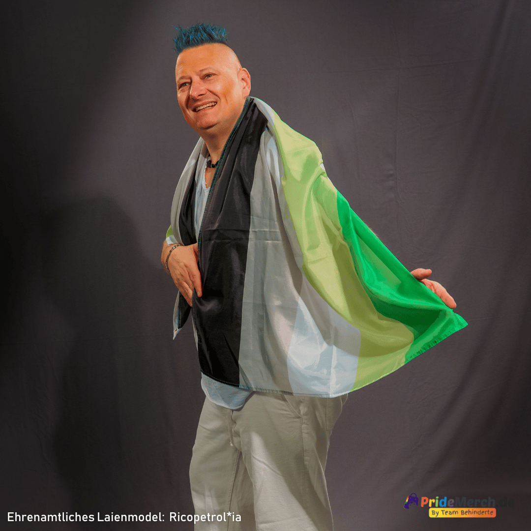 Das ehrenamtliche Laienmodel Ricopetrolia (bekannt als Bremer Drag Queen Gina Petrolia) präsentiert gut gelaunt im Profil die große Aromantische Pride Flagge im Format 150x90 cm. Das Foto zeigt das Model mit blau gefärbtem Irokesenschnitt, wie es sich die Flagge mit den Farben Dunkelgrün, Hellgrün, Weiß, Grau und Schwarz stolz wie einen Umhang über die Schulter gelegt hat. Unten rechts ist das Logo von PrideMerch.de zu sehen.