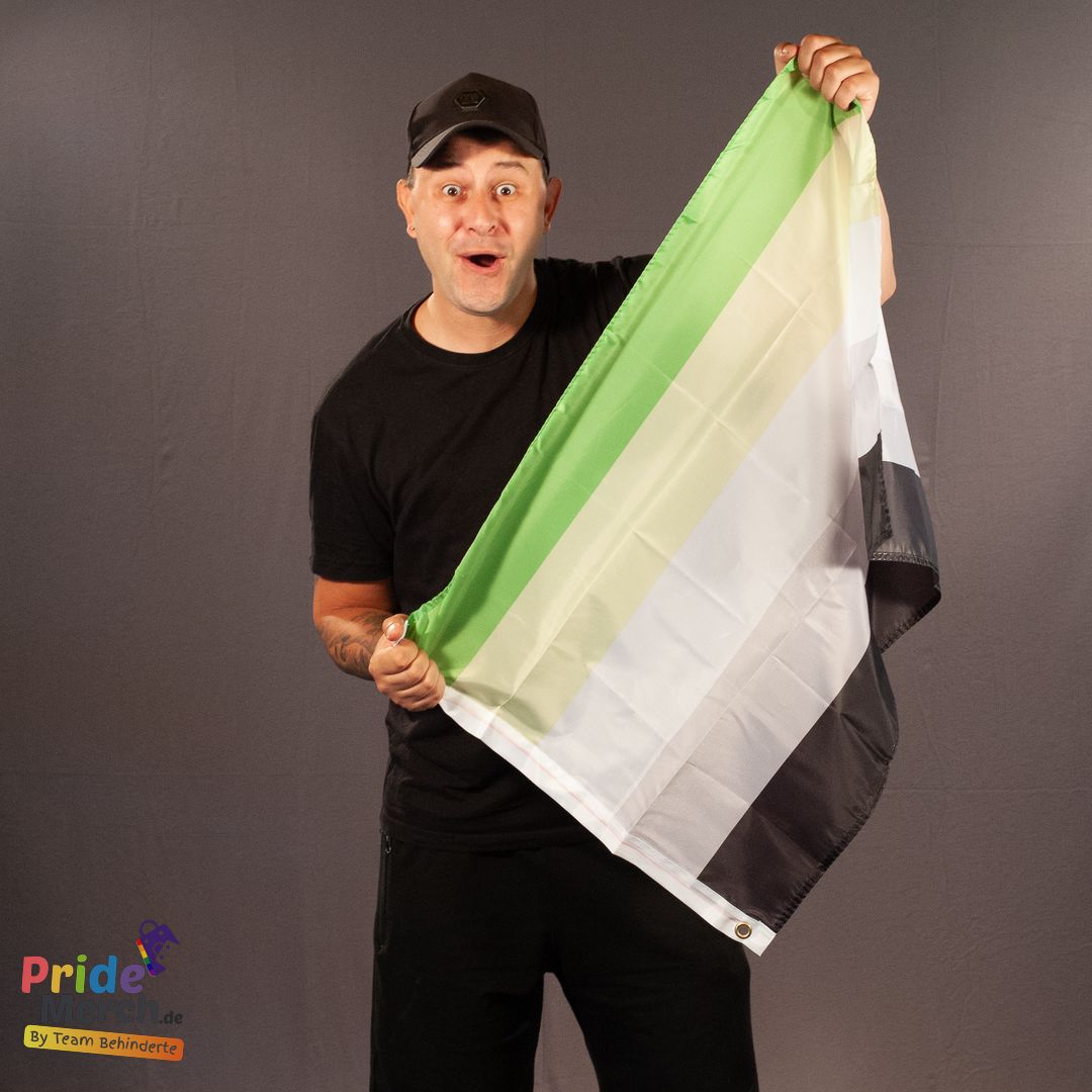 Auf dem Studiofoto präsentiert Bremens bekannteste Drag Queen, Joyce DeLone, mit einem herrlich überraschten Gesichtsausdruck die kleinere Variante der aromantischen Flagge im Format 90x60 cm. Bekleidet mit schwarzem T-Shirt und schwarzer Basecap hält Joyce die Flagge dynamisch hoch, sodass die gut sichtbare Metallöse an der unteren weißen Kante auffällt. Unten links befindet sich das Logo von PrideMerch.de.