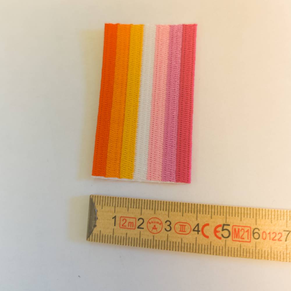 Warme Orangetöne, ein mittiger weißer Streifen und kräftige Pinknuancen bilden das längsgestreifte Muster dieses Lesbian Pride Aufnähers. Das gestickte Rechteck ist ringsum mit einem weißen Faden eingefasst. Ein gelber Zollstock direkt darunter dient als Größenvergleich und markiert eine exakte Breite von dreieinhalb Zentimetern.