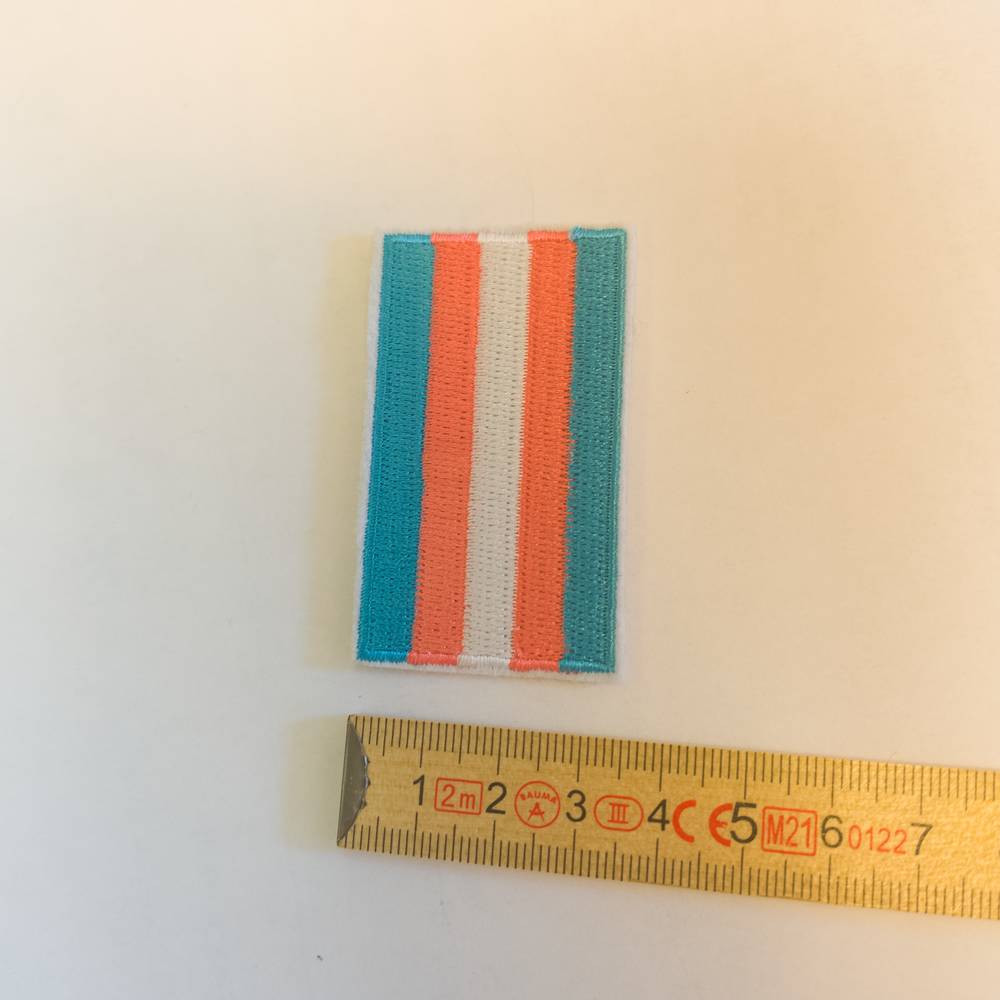 Ein kleiner, fein gestickter Stoff-Patch in den Farben der Trans-Pride. Die hellblauen, rosafarbenen und weißen Streifen verlaufen hier von links nach rechts, eingefasst von einem schmalen weißen Rand. Zur räumlichen Orientierung liegt der Aufnäher auf einem hellen Untergrund direkt über einem gelben Holzzollstock, der eine Breite von knapp dreieinhalb Zentimetern anzeigt.