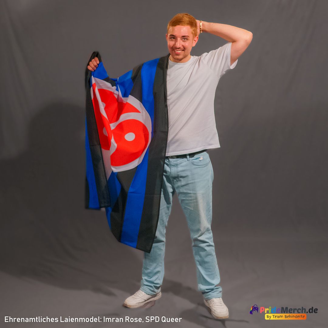 Imran Rose steht frontal und breit lächelnd vor einem grauen Hintergrund. Er trägt ein weißes T-Shirt und eine hellblaue Jeans. Seine linke Hand liegt entspannt am Hinterkopf. Mit der rechten Hand hält er die BDSM-Flagge hoch, die über seine rechte Schulter drapiert ist und locker an seiner Seite herabfällt. Das rote Triskelion und die dunklen, klaren Streifen der Flagge bilden einen starken optischen Kontrast zu seinem hellen Outfit.