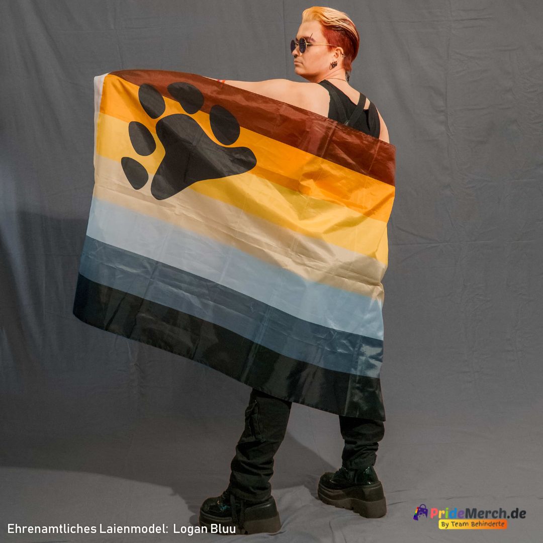 Der Bremer Drag King Logan Bluu steht mit dem Rücken zur Kamera vor einem grauen Hintergrund und blickt cool über seine rechte Schulter. Er hat rötlich-blondes, hochgestyltes Haar und trägt eine runde Sonnenbrille, ein schwarzes Oberteil, eine schwarze Cargohose sowie klobige schwarze Plateau-Boots. Die Bear Pride Flagge ist lässig über seinen Rücken und seine Schultern drapiert. Die schwarze Bärentatze prangt deutlich auf seinem linken Schulterblatt.