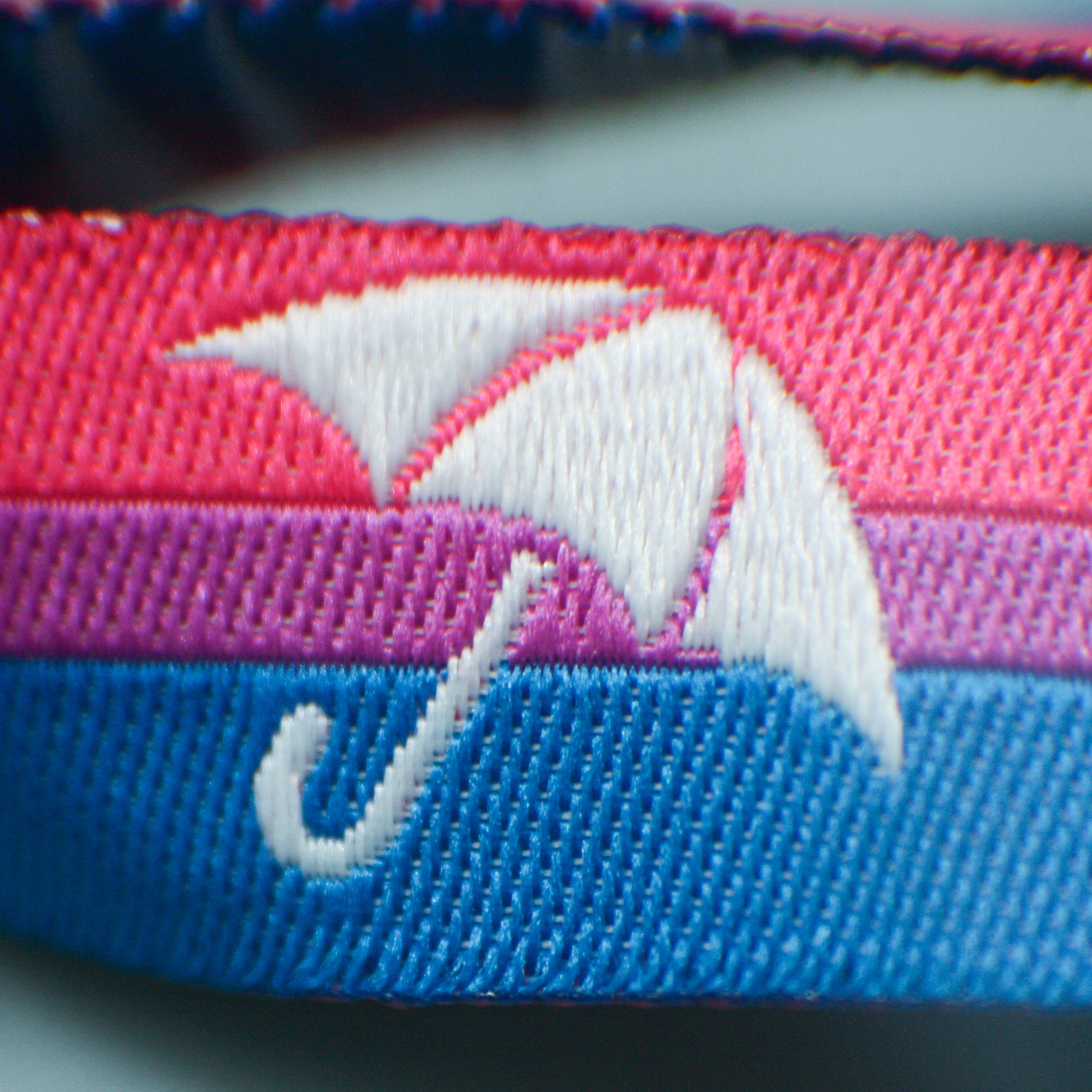 Eine extreme Makro-Nahaufnahme des Bi+ Pride Stoffarmbändchens. Im Fokus steht das detailliert eingewebte, weiße Regenschirm-Symbol des Partnervereins Bi+ Hamburg e.V. Die feine Struktur des gewebten Bandes zeigt die drei horizontalen Streifen in den Bisexual-Pride-Farben Pink, Lila und Blau. Dieses Solidaritätsprodukt unterstützt das Inklusionsprojekt Team Behinderte.
