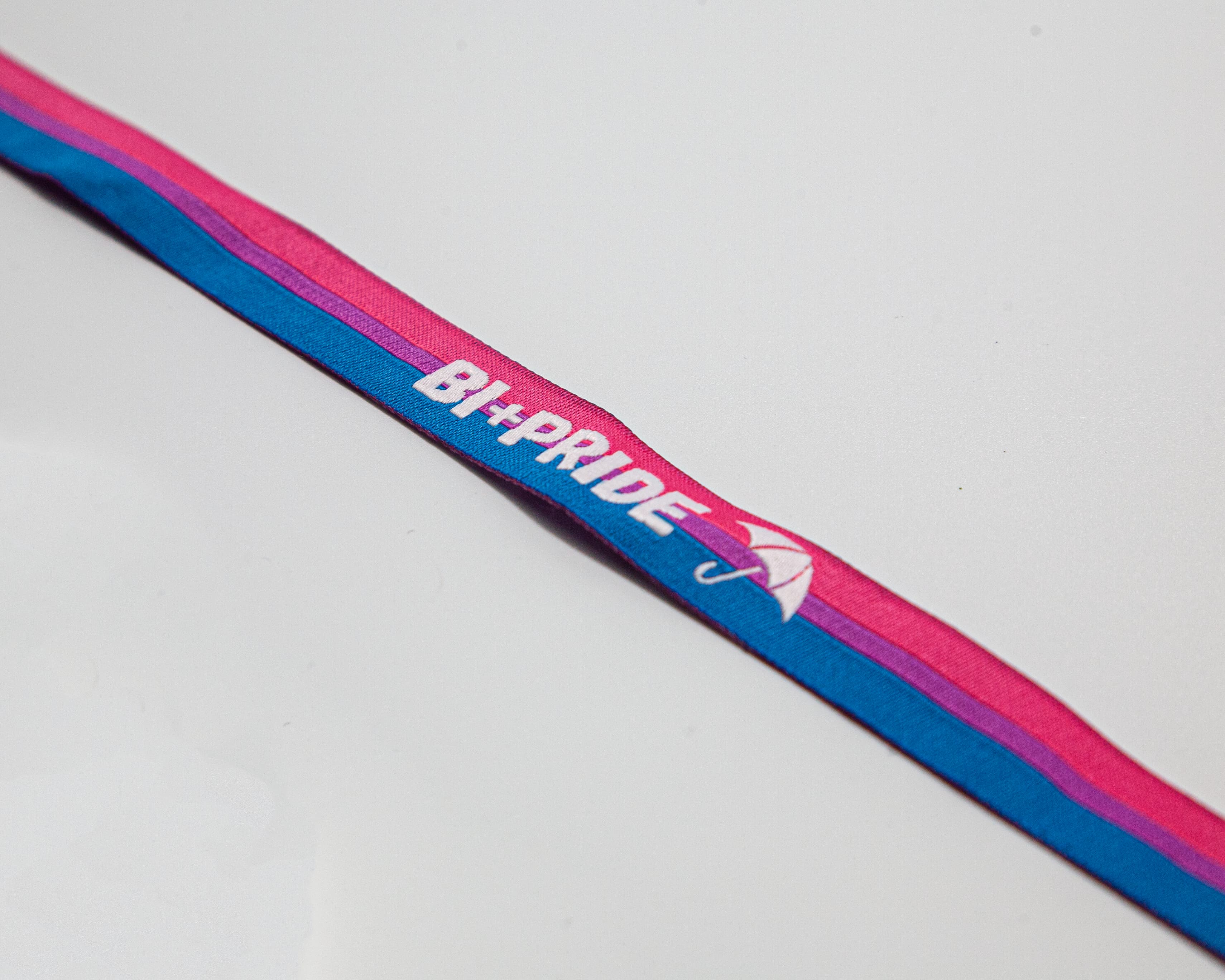 Ein hochwertig gewebtes Festivalbändchen in den Bi-Pride-Farben Pink, Lila und Blau, das elegant in Herzform auf weißem Untergrund gelegt ist. Deutlich sichtbar ist der eingestickte weiße Schriftzug „BI+PRIDE“ und das Regenschirm-Symbol des Partnervereins. Am linken Ende befindet sich der stufenlos verstellbare Metallverschluss. Ein Kauf unterstützt mit 10 % Umsatzbeteiligung die Bi+ Sichtbarkeit in Hamburg.