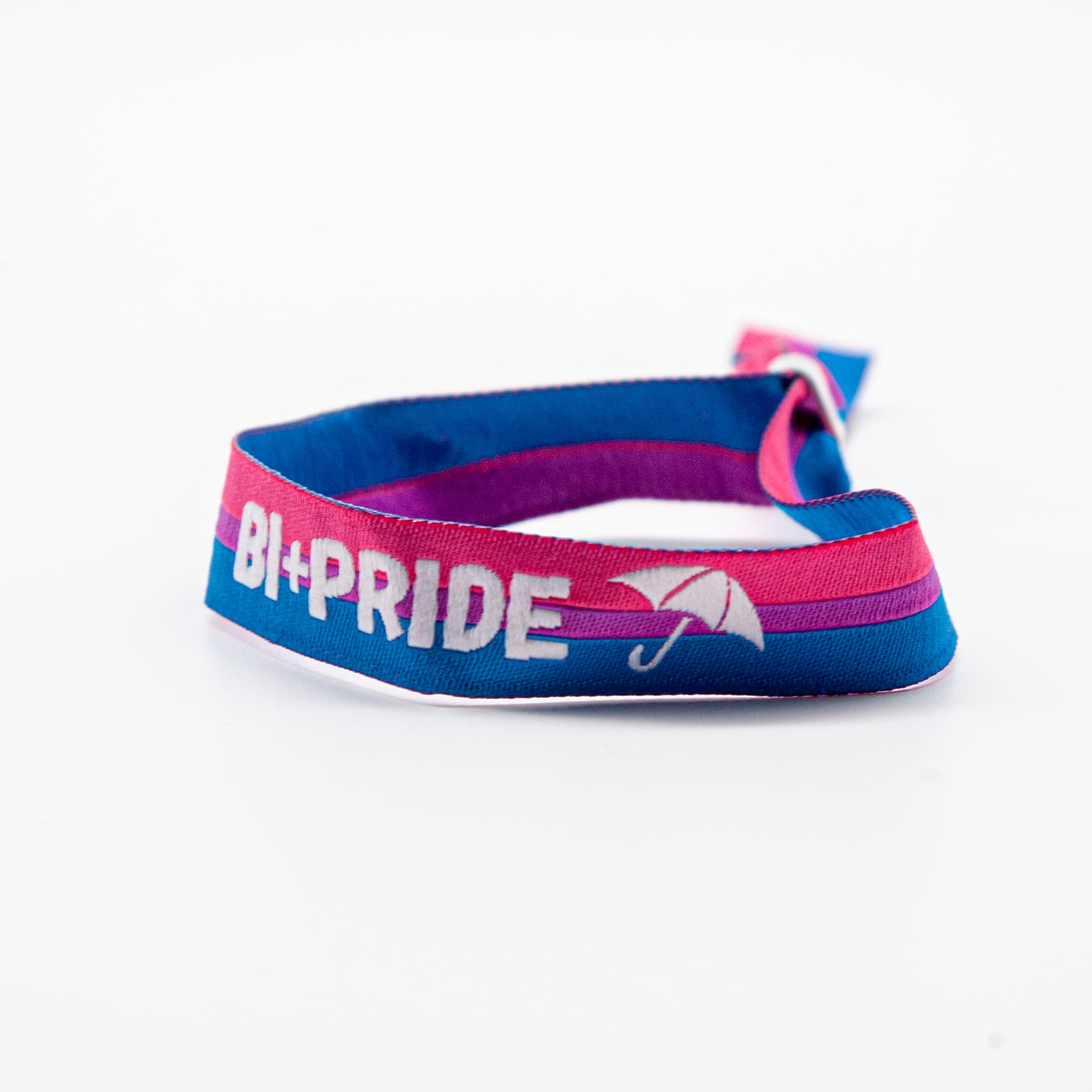 Ein geschlossenes, rund aufgestelltes Festival-Stoffarmband in den Farben der Bisexual Pride Flagge (Pink, Lila, Blau). Gut sichtbar ist die weiße Einstickung 'BI+PRIDE' mit dem Schirm-Symbol auf weißem Grund. Mit diesem Solidaritätsarmband werden 10% des Umsatzes an den Bi+ Hamburg e.V. gespendet, um queere Sichtbarkeit im Norden nachhaltig zu fördern.