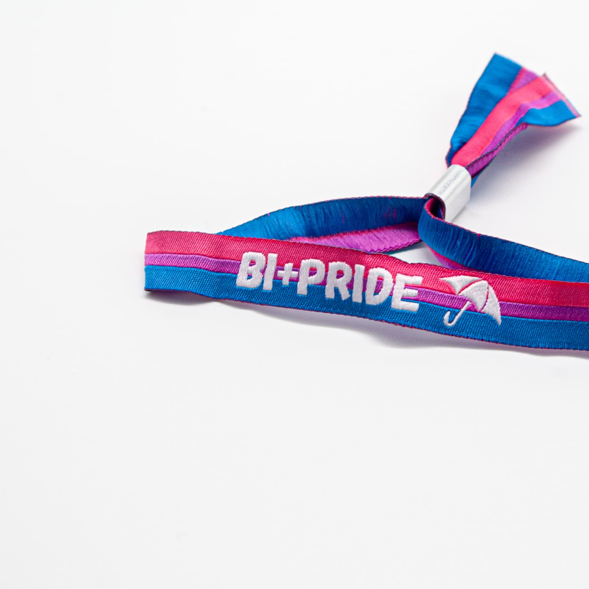 Nahaufnahme eines gewebten Festivalbändchens für die Bi+ Pride Hamburg. Der weiße Schriftzug 'BI+PRIDE' und das Regenschirm-Logo sind sauber auf dem pink-lila-blauen Stoff eingestickt. Am Ende befindet sich ein silberner Metallverschluss. Ein Kauf dieses Partnerprodukts sichert Arbeitsplätze für queere Menschen mit Behinderungen abseits von Werkstätten.