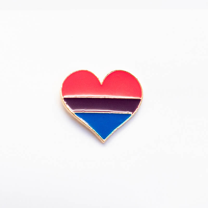 Auf einer hellgrauen Holzstruktur ist die bisexuelle Variante des Emaille-Herz-Pins flach liegend abgebildet. Das Schmuckstück präsentiert die unmissverständliche Farbkombination aus Pink, einem schmalen lila Mittelstreifen und Dunkelblau.