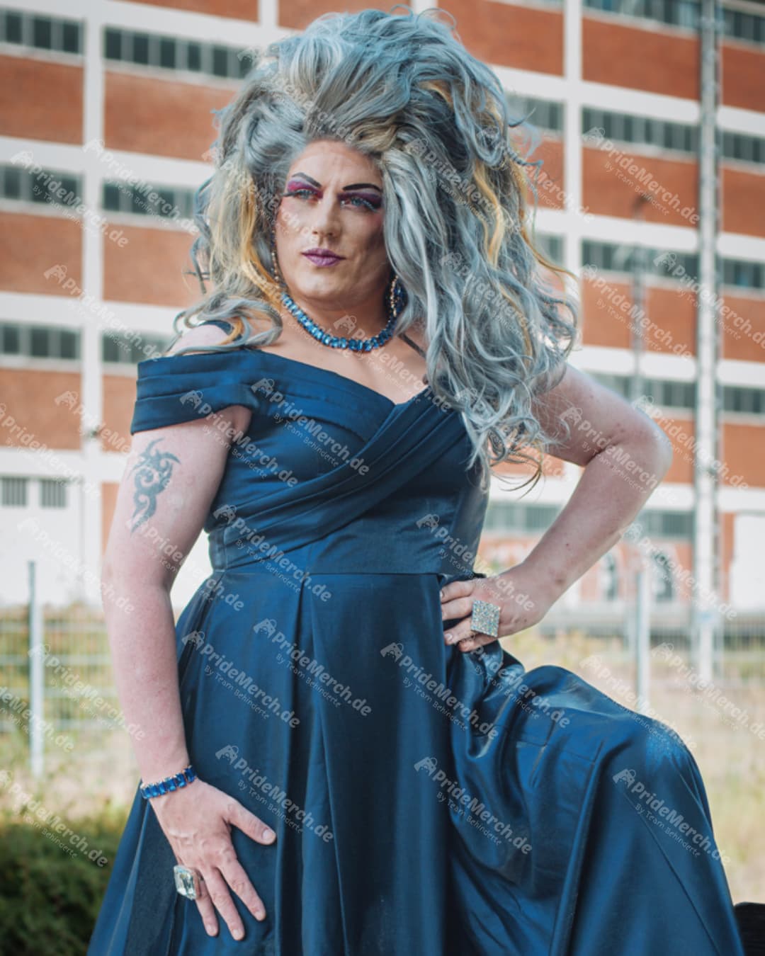 Porträt von Jolina Wings, Mitglied von "Drag Queens & Drag Kings on PrideMerch.de", in einer Power-Pose mit Hand in der Hüfte, dramatischem Augen-Make-up und blauem Schmuck blickt selbstsicher zur Seite.
