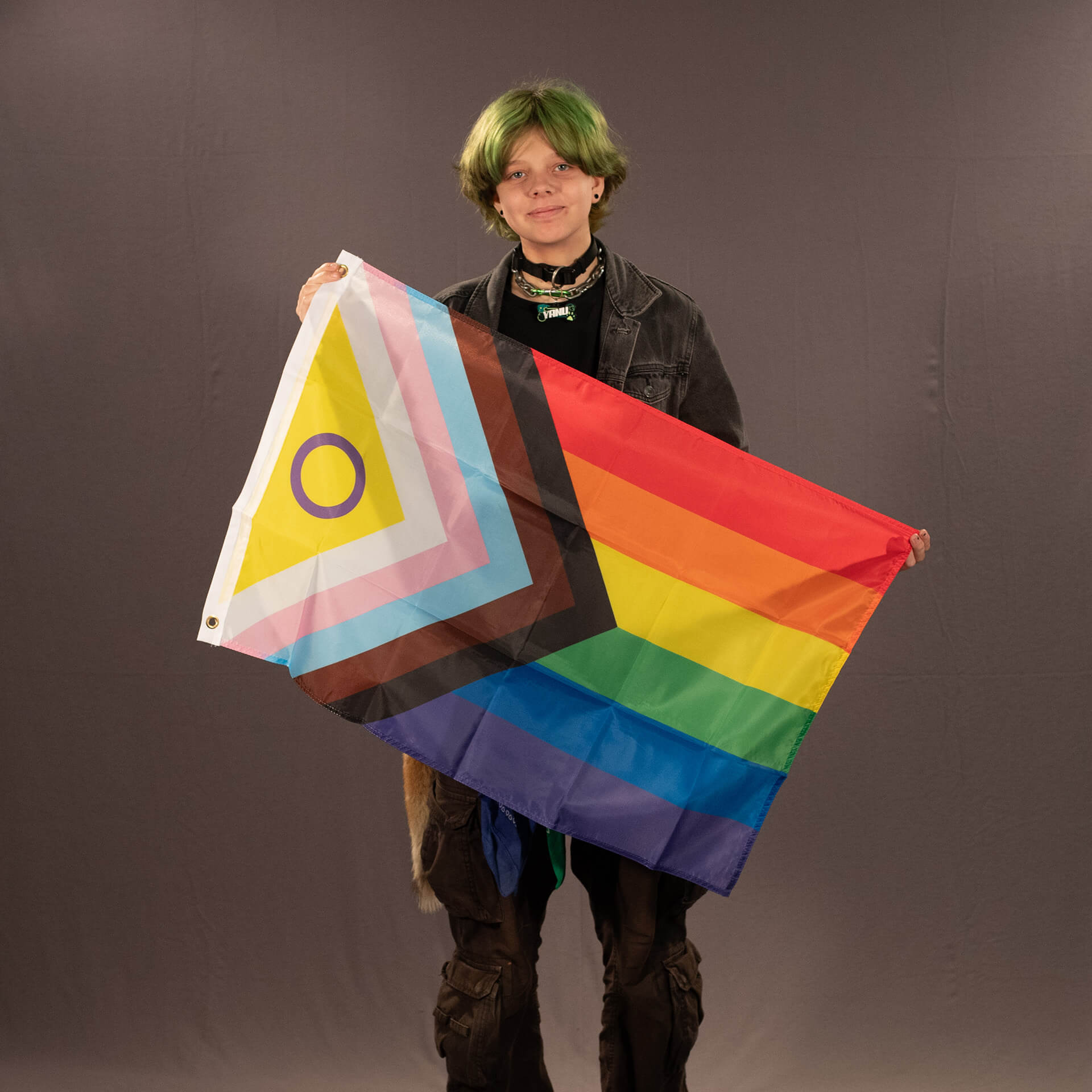 Das ehrenamtliche Laienmodell Chris präsentiert die kompakte, 90x60 cm große Intersex Inclusive Progress Pride Flagge vor einem grauen Studiohintergrund. Er hält das farbintensive Textil diagonal vor sich, wodurch der weiße Rand mit den goldfarbenen Befestigungsösen links oben gut sichtbar wird.