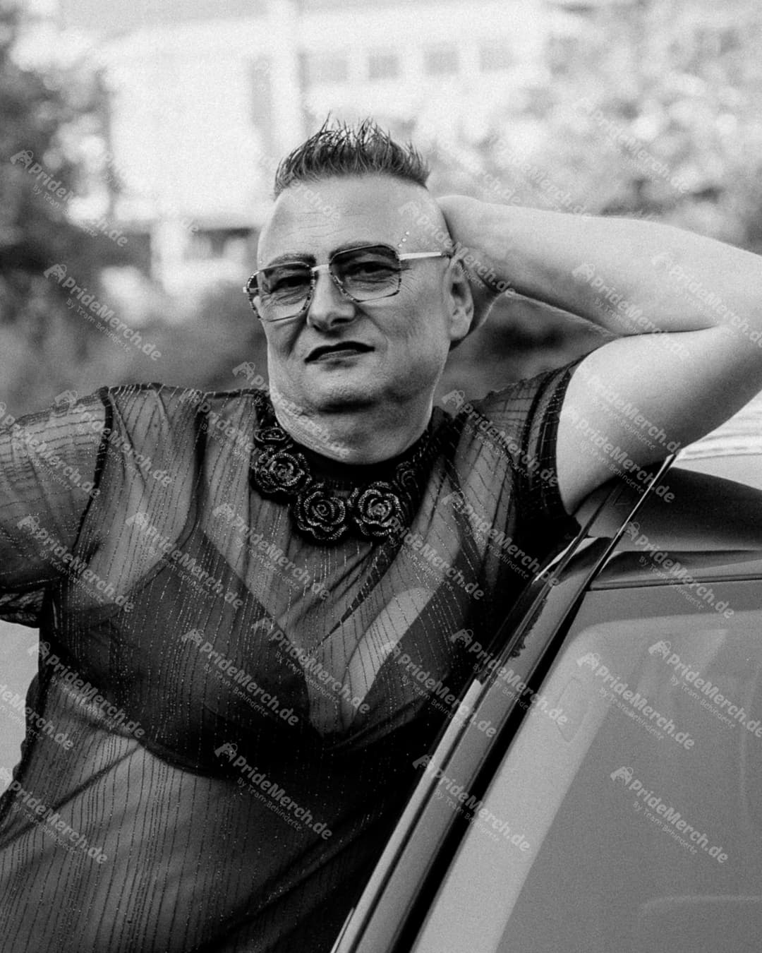 Die Drag-Künstlerin Gina Petrolia in einem transparenten, gestreiften Oberteil und mit markanter Brille blickt für PrideMerch.de selbstsicher in die Kamera.