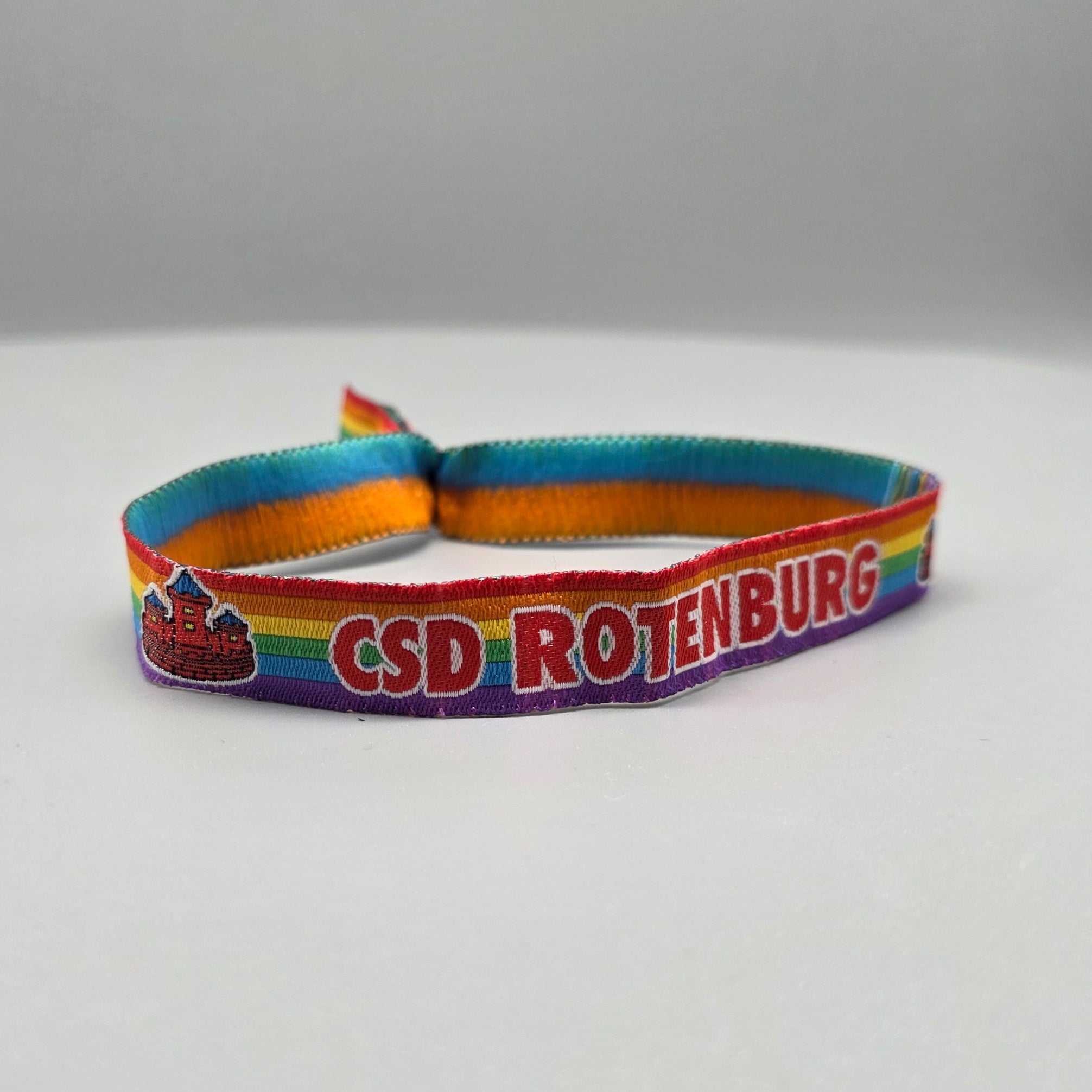 Ein Regenbogen-Stoffarmband für den CSD Rotenburg, ausgestattet mit einer silbernen Metallplombe. Der rote Schriftzug „CSD ROTENBURG“ mit weißer Kontur wird von einem detaillierten roten Burg-Motiv flankiert und setzt ein klares Zeichen für lokalen Aktivismus.
