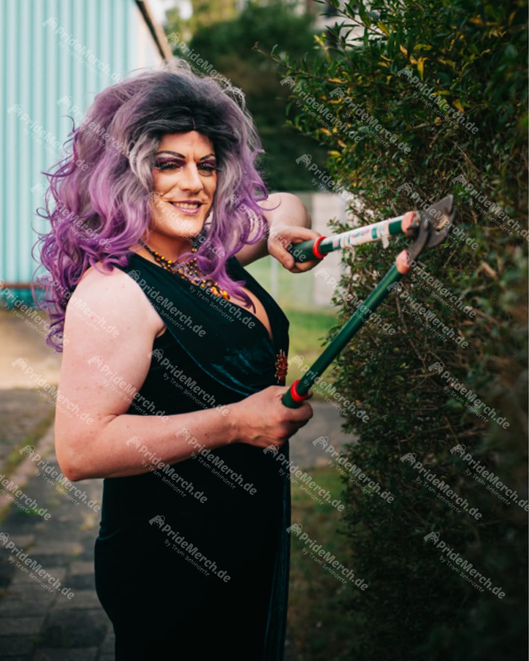 Die Drag Queen Jolina Wings in einem eleganten schwarzen Samtkleid und mit einer großen lila Perücke lächelt, während sie mit einer Heckenschere einen Busch schneidet.