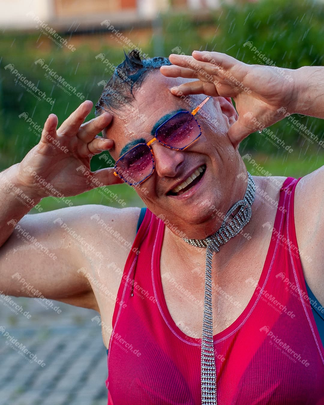 Die Drag-Künstlerin Gina Petrolia in einem pinken Trägertop und mit einer silbernen Krawatten-Halskette blickt lachend in die Kamera, während es auf sie regnet