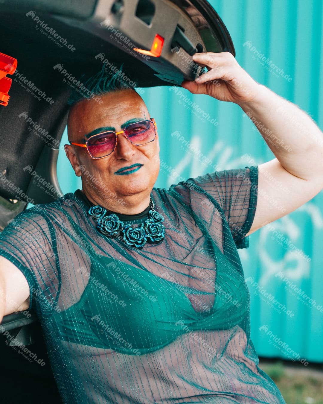 Comedy Drag Queen Gina Petrolia posiert selbstbewusst im Kofferraum eines Autos in einem kompletten türkisfarbenen Look mit Mesh-Oberteil und Sonnenbrille