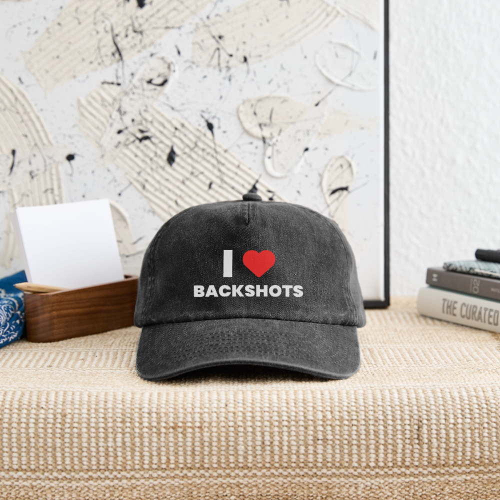 Die verwaschene schwarze Schirmmütze liegt griffbereit auf einer Ablage neben dem Eingang. Das kinky „I ❤️ BACKSHOTS“-Design macht das Teil zum idealen, sex-positiven Begleiter für die nächste Festival- und Clubnacht.