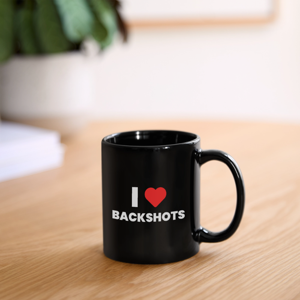 Die schwarze Kinky-Kaffeetasse steht einsatzbereit auf einem modern eingerichteten Schreibtisch neben einer Tastatur. Das farbintensive rote Herz mit dem weißen Text „I ❤️ BACKSHOTS“ setzt einen humorvollen, sex-positiven Akzent im Arbeitsalltag.