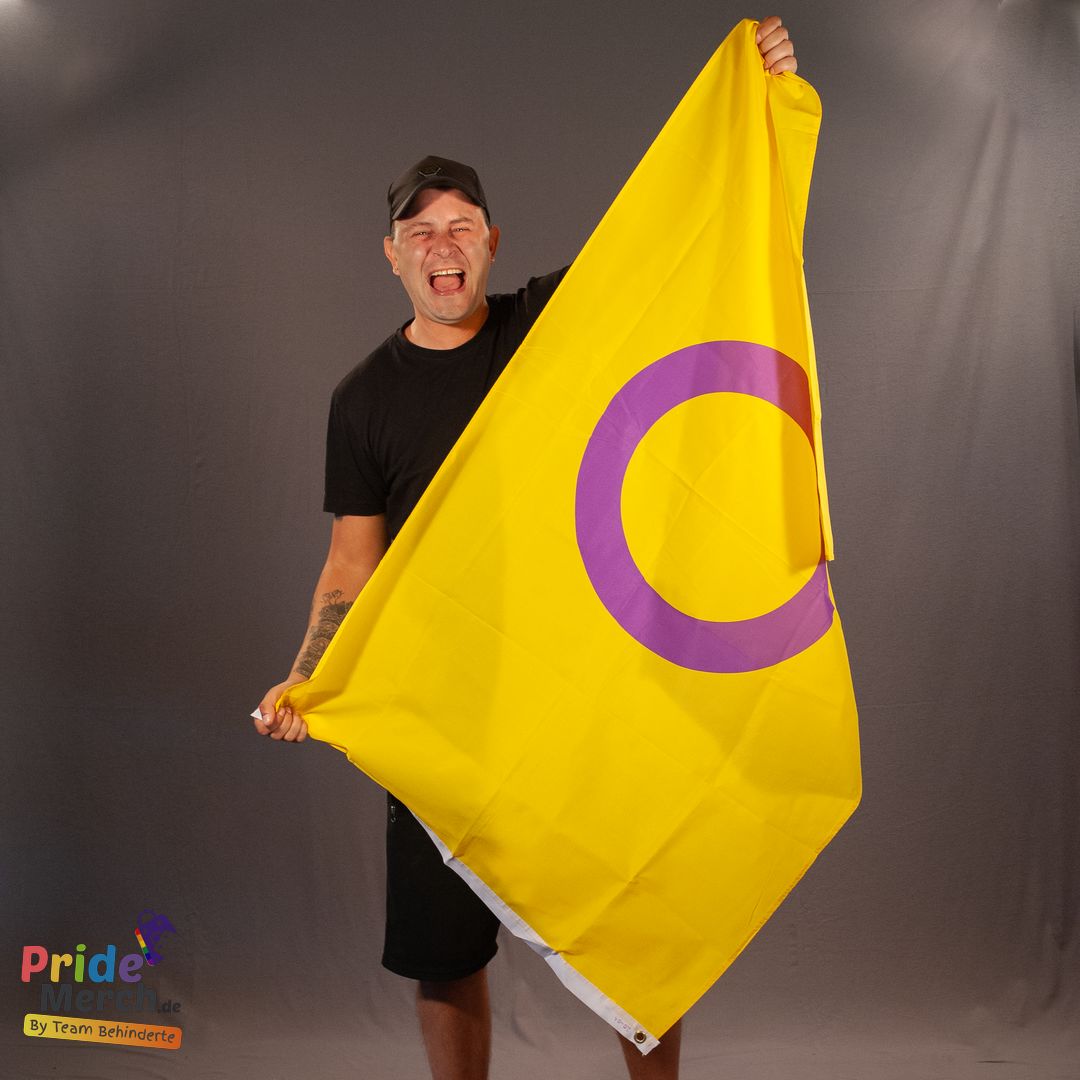 Joyce DeLone (Out of Drag) präsentiert die Intersex-Flagge mit einem freudig geöffneten Mund und sichtbarer Begeisterung. Joyce steht vor einem grauen Hintergrund, trägt eine schwarze Baseballkappe, ein schlichtes schwarzes T-Shirt und schwarze Shorts. Auf dem rechten Unterarm sind Tattoos zu erkennen. Die gelbe Flagge mit dem lila Ring wird diagonal vor dem Körper aufgespannt, sodass das zentrale Kreismotiv deutlich sichtbar ist.
