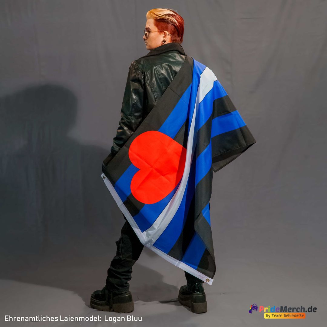 Das ehrenamtliche Laienmodell Logan Bluu ist von hinten schräg im Profil zu sehen und blickt cool über die linke Schulter zurück. Logan hat kurze Haare mit blondem Deckhaar und rötlich-dunklen Seiten, trägt eine runde Sonnenbrille, eine schwarze Lederjacke, dunkle Hosen und markante schwarze Plateau-Schuhe. Die Leather Pride Flagge ist wie ein Umhang lässig über die rechte Schulter und den Rücken geworfen. Das große rote Herz der Fahne leuchtet dabei auffällig auf Kniehöhe.