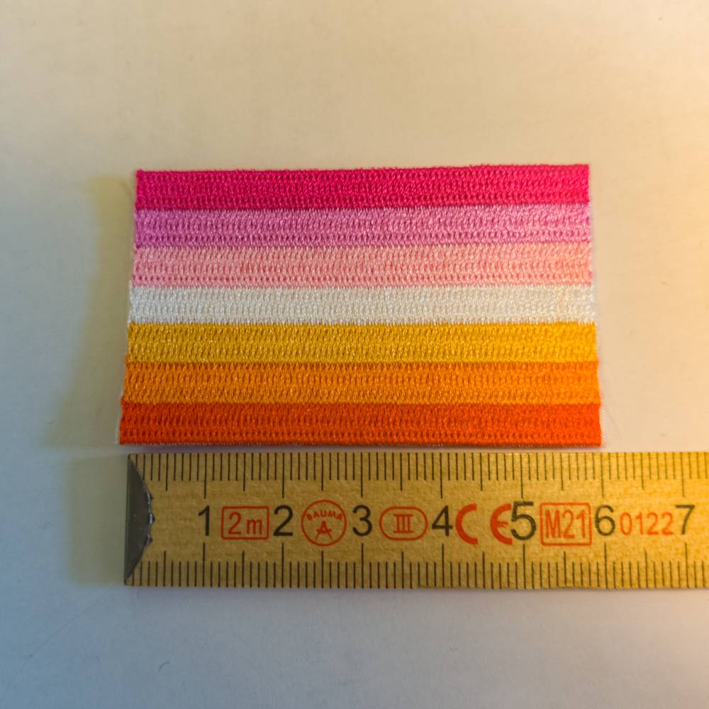 Ein Textil-Aufnäher, der komplett aus glänzendem Stickgarn besteht. Er zeigt die sieben horizontalen Streifen der Lesbian Sunset Flagge in kräftigen Pink-, Rosa-, Weiß- und Orangetönen. Zur exakten Größeneinordnung liegt der Patch direkt oberhalb der Zentimeter-Skala eines gelben Holz-Zollstocks.
