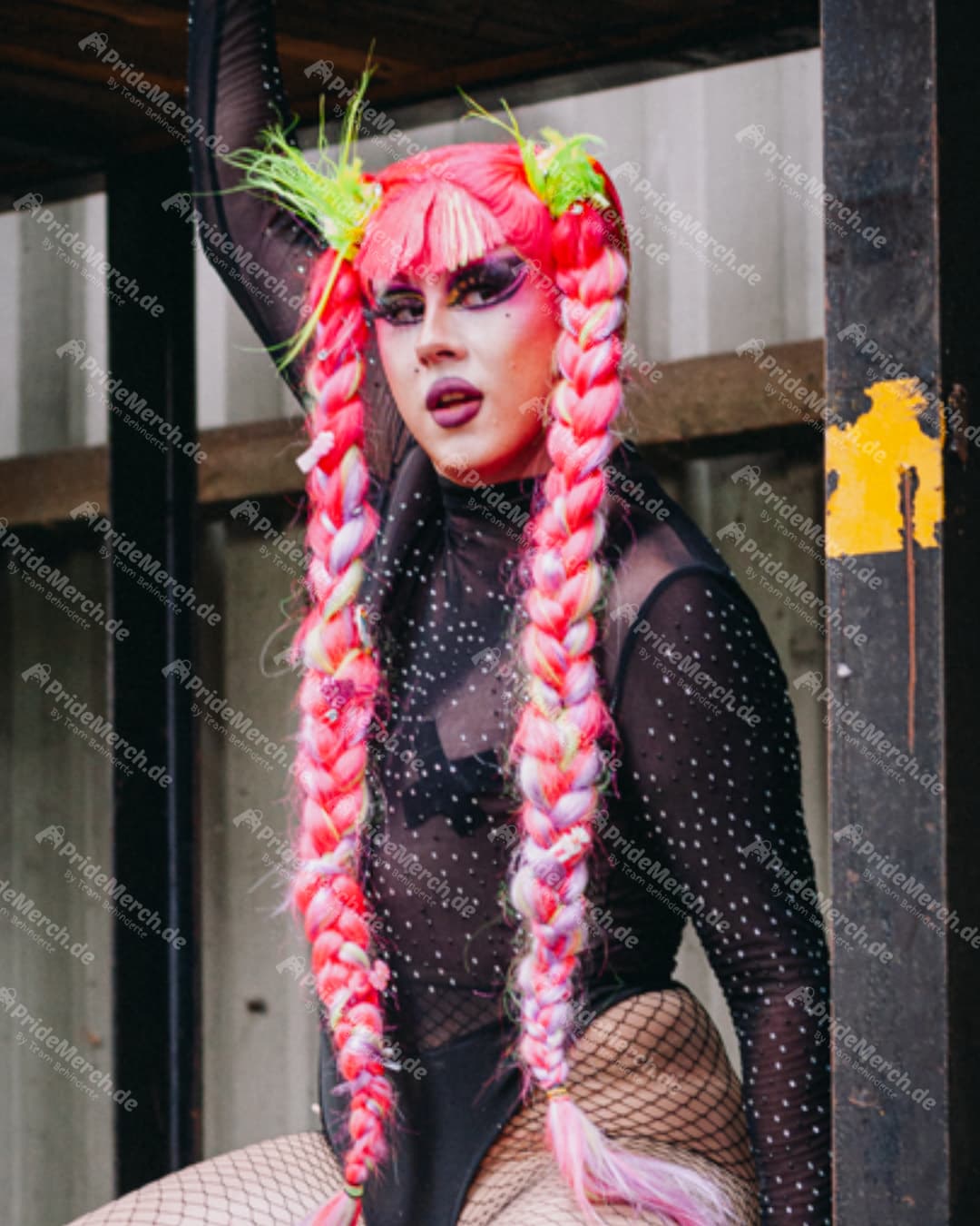 Drag-Performer mit neonpinken Flechtzöpfen, lila Augen-Make-up und schwarzem Mesh-Body in industriellem Umfeld