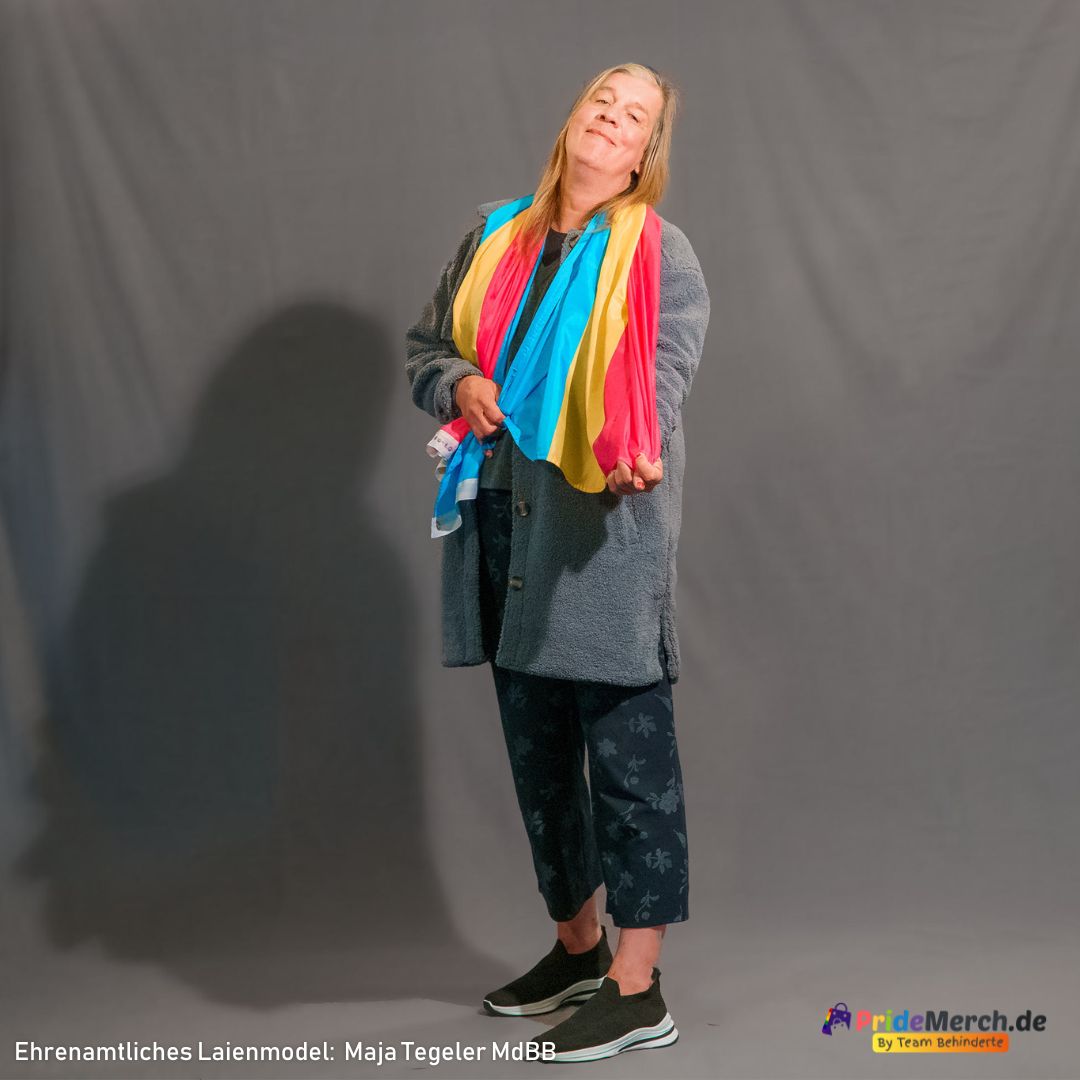 Das ehrenamtliche Laienmodell Maja Tegeler steht vor einem grauen Fotohintergrund. Sie hat langes blondes Haar, lächelt entspannt und trägt eine dunkelgraue, flauschige Jacke über einem dunklen Oberteil sowie eine bläuliche Hose mit dezentem Muster und schwarze Sneaker. Die große Pansexuell Pride Flagge in den Farben Pink, Gelb und Blau hat sie sich elegant wie einen bunten Schal eng um den Hals und die Schultern gelegt. Sie hält die Enden der Flagge locker mit den Händen auf Bauchhöhe.