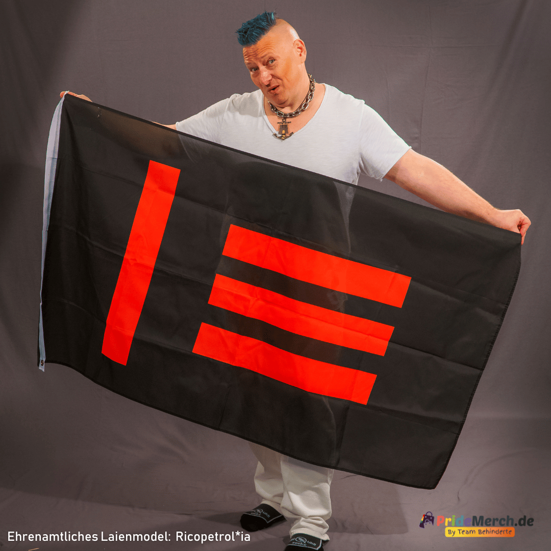Laienmodell Gina Petrolia (Ricopetrol*ia) posiert lässig vor einem grauen Hintergrund und hält die schwarz-rote Master/Slave-Flagge weit geöffnet. Gina trägt einen auffälligen blauen Irokesenschnitt, ein weißes T-Shirt mit V-Ausschnitt, eine massive silberne Gliederkette mit Anker-Anhänger, eine weiße Hose und dunkle Socken mit weißen Badelatschen. Die asymmetrische, rote Linien-Grafik der Flagge ist klar und deutlich auf dem schwarzen Stoff zu erkennen.