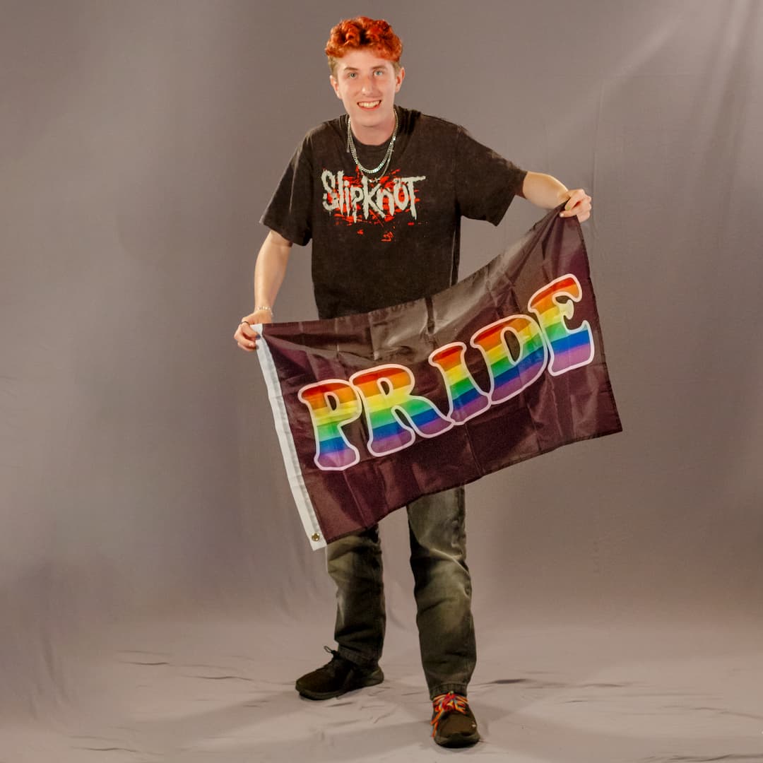 Das ehrenamtliche Laienmodell hält lächelnd die schwarze Flagge mit dem farbenfrohen PRIDE-Schriftzug schräg vor sich. Die Studioaufnahme zeigt das leichte, wehende Polyester-Material der kompakten Flaggenvariante.