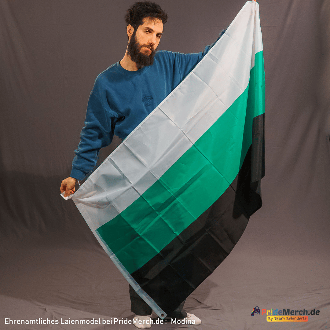 Das ehrenamtliche Modell Modina, ein Mann mit dunklem Vollbart und kurzem Haar, trägt einen blauen Pullover. Er hält die Neutrois-Flagge im Fotostudio schräg vor sich ausgebreitet. Die drei horizontalen Streifen der Flagge – oben Weiß, in der Mitte ein leuchtendes Grün und unten tiefes Schwarz – sind durch die Bewegung des Stoffes dynamisch dargestellt. Das Bild zeigt die Größe von 150x90 cm und die kräftige Farbwirkung des neutral-gender Designs.