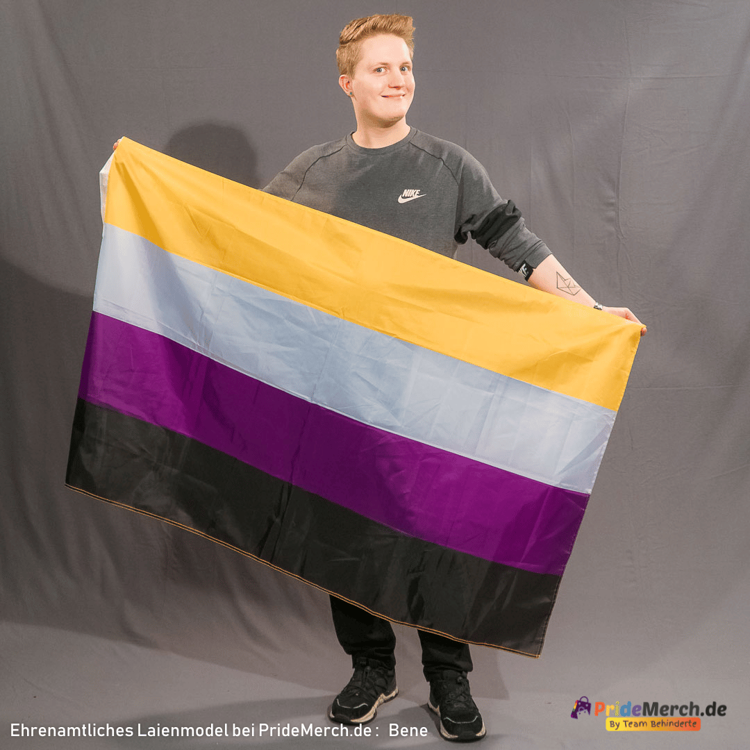 Bene steht entspannt vor einem grauen Hintergrund und lächelt sanft in die Linse. Er hält die große Non-Binary Flagge an den oberen Ecken weit ausgebreitet vor seinem Körper, wodurch die klaren Farben Gelb, Weiß, Lila und Schwarz vollständig und faltenfrei sichtbar sind.