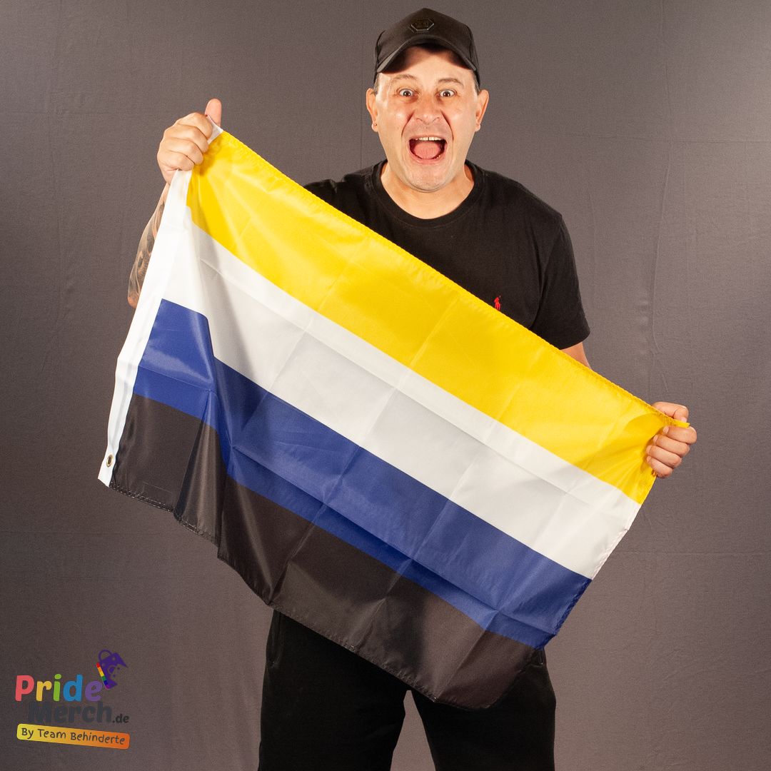 Joyce DeLone (out of Drag) steht vor einem grauen Hintergrund und reißt den Mund zu einem begeisterten Jubeln auf. Er trägt ein schwarzes T-Shirt und eine schwarze Cap. Mit beiden Händen präsentiert er die kleinere 90x60 cm Version der Non-Binary Flagge straff vor seiner Brust.