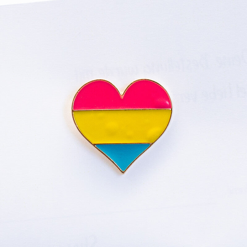 Zwei glänzende, silberne Metalllinien unterteilen diesen herzförmigen Pride-Anstecker in die drei offiziellen Farben der pansexuellen Flagge. Die intensiven Bahnen in Pink, Gelb und Blau heben sich leuchtend von der hellen Unterlage ab.