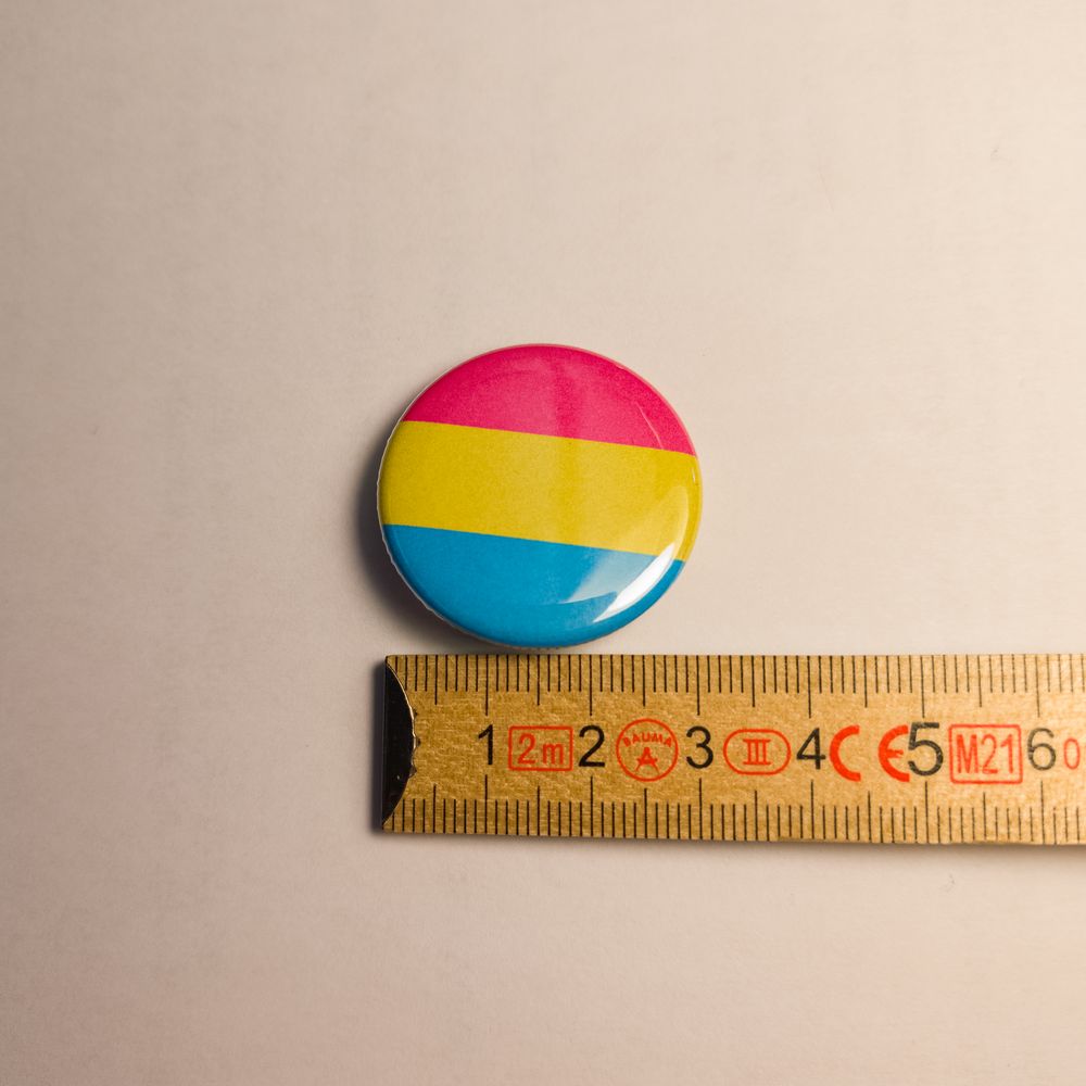 Ein runder, glänzender Metall-Button zeigt die pansexuelle Flagge mit ihren drei Streifen in Pink, Gelb und Blau. Der Pin liegt bündig an einem Zollstock an, welcher einen Durchmesser von knapp 31 Millimetern verdeutlicht.