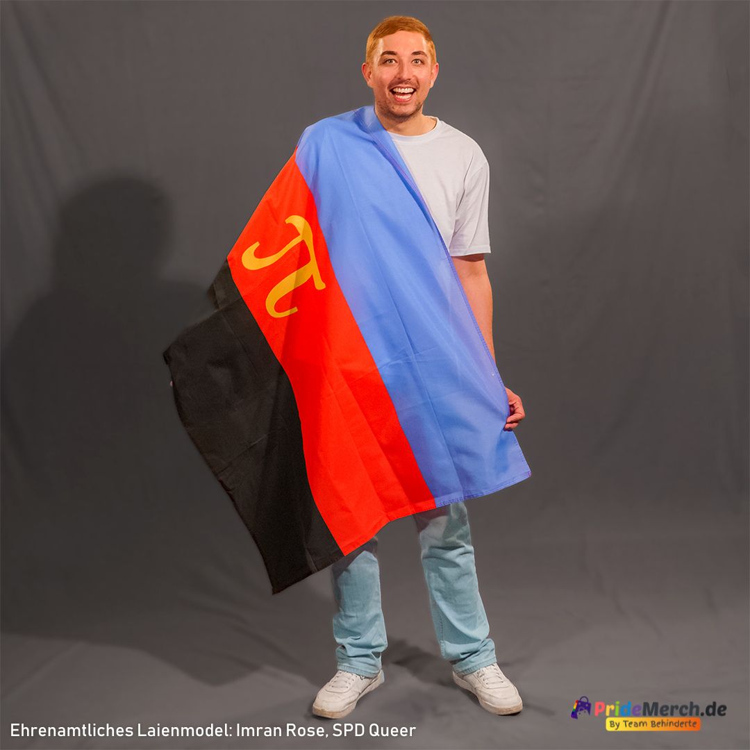 Imran Rose lacht fröhlich und unbeschwert in die Kamera. Er hat die klassische Polyamorie Flagge über seine rechte Schulter geworfen und hält sie vor seinem Körper zusammen, sodass der markante rote Streifen mit dem gelben Pi-Symbol und der hellblaue Streifen im Fokus stehen.